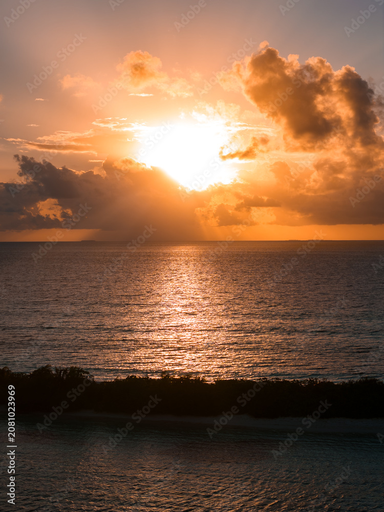 Fototapeta premium Sun breaks through towering cumulus clouds near Dhigurah Island, Maldives, casting a golden path on calm water, with a thin island silhouette and soft reflections.