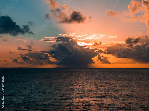 Sun sets near Dhigurah Island in the Maldives, with anvil clouds glowing orange and pink. Calm sea reflects teal and copper tones, suggesting a warm tropical dusk.