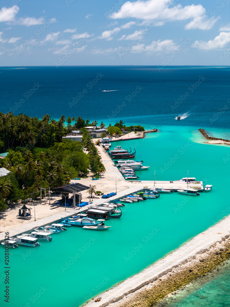 Fototapeta premium Aerial midday view of Dhigurah Island, Maldives, with slim sand and coral shore, compact marina, palms, narrow road, two boats leaving wakes toward the Indian Ocean.