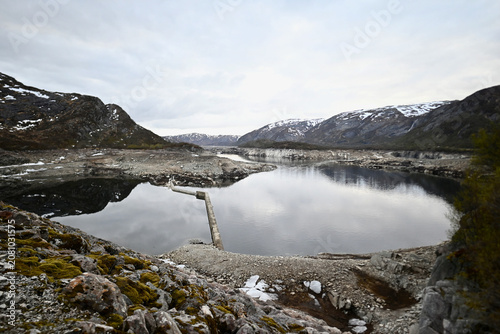 Reservoir above a dam in Norway, surrounded by mountains lightly covered with snow, under a calm overcast evening sky in late spring.