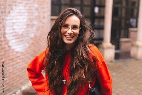 Portrait beautiful young brunette woman with long hair wearing gold-rimmed glasses and a vibrant red oversized sweatshirt laughing heartily looking at camera in a stylish urban alleyway street style
