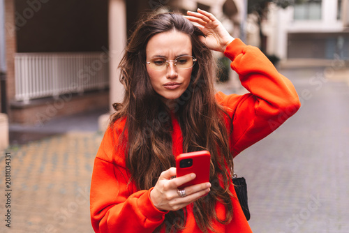 Frustrated young woman looking at smartphone screen with an annoyed and puzzled expression while touching her hair in an urban setting wearing an orange sweatshirt and yellow tinted eyeglasses