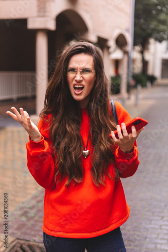 Obraz na plátně Furious young woman shouting and gesturing in anger while holding a smartphone o