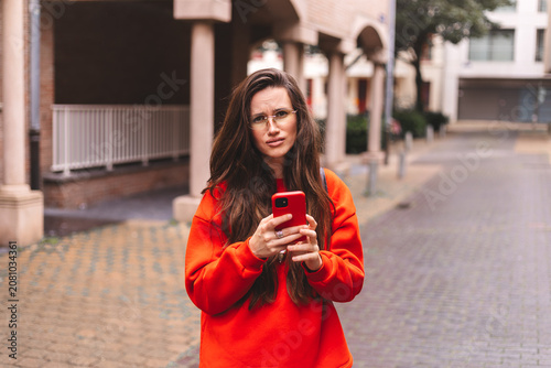 Skeptical young woman with long wavy hair wearing an orange sweatshirt and yellow eyeglasses holding a smartphone and looking directly at the camera with an expression of doubt and judgment