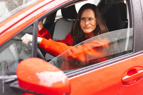 Disgusted woman with long brunette hair in orange sweatshirt and eyeglasses driving red car in urban city setting, side view through open window, expressing annoyance, disapproval skeptical grimace