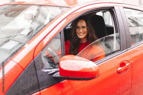 Smiling woman with long brunette hair wearing orange sweatshirt and eyeglasses driving red car in urban city setting, side view through open window, friendly and confident look, authentic joy portrait