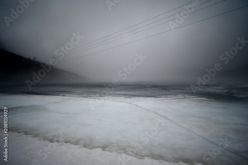 Frozen mountain lake in Norway with a small patch of open water, surrounded by mist and rugged peaks during a foggy late autumn day.