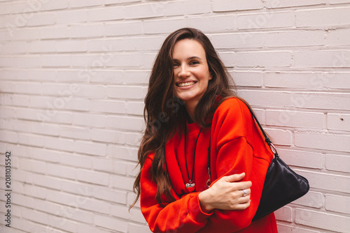 Cuadro en lienzo Laughing young woman in red sweatshirt looking at camera while leaning against w