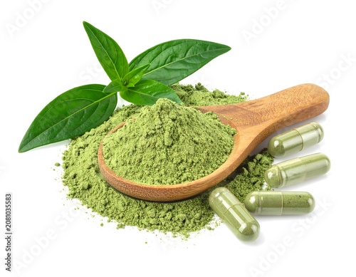 A close-up shot of green herbal powder spilling from a wooden spoon, fresh leaves, and capsules