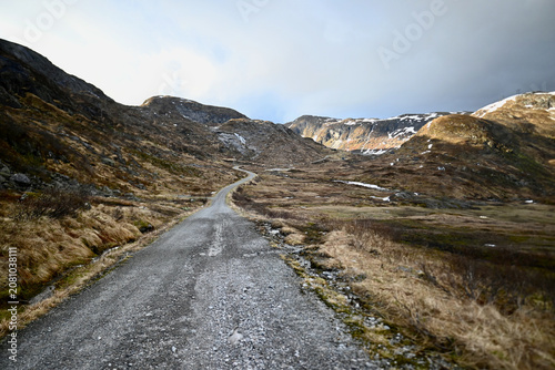 Canvas Print Gravel road stretching into the distance through the mountains of Norway in late spring, surrounded by dry grass and small patches of lingering snow