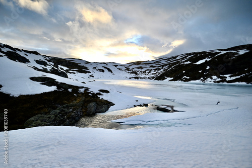 Frozen mountain lake in Norway glowing in sunset light, with melting ice revealing open water amid snowy peaks during late spring.