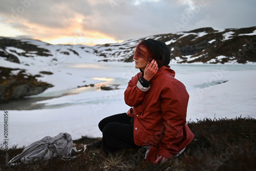Woman in an orange rain jacket sitting by a snowy mountain lake in Norway at sunset, holding her hair in strong wind with dramatic alpine peaks behind.