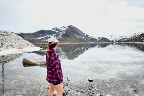 Woman in a pink-purple shirt and cap standing with her back to the camera by a mountain lake in Norway in May, with lightly snow-covered peaks behind.