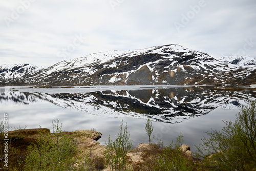 Mountain lake above a Norwegian fjord with mirror reflections of peaks lightly covered in snow during late spring.
