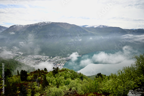 Panoramic view of Høyanger village in Norway from above the fjord, with turquoise water, a scenic valley, and low clouds drifting over the landscape.