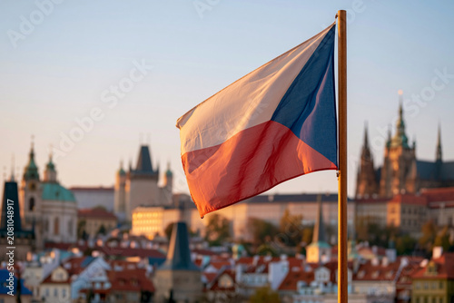 Czech flag waves gently in the breeze. Historic city skyline rises behind it. Golden hour bathes buildings in warm light. Red, white, and blue colors stand out vividly. Ideal for travel, cultural