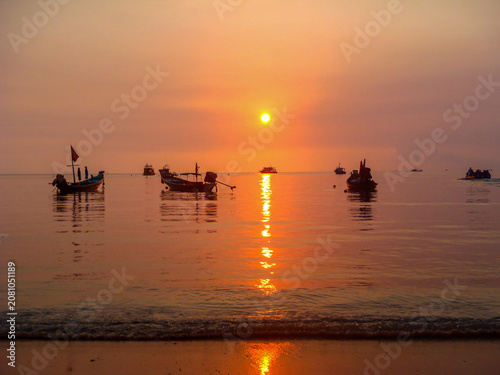 Hazy orange sunset with reflections over calm bay with boats in Thailand