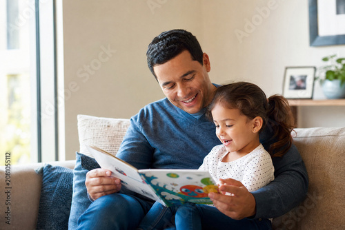 Pai lendo livro ilustrado para sua filha pequena em um sofá aconchegante - Conceito de educação, carinho paterno e tempo de qualidade em família.