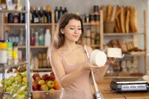 Surrounded by warm, homely ambiance of small grocery shop, young woman holding up packages artisan cheeses, comparing and contemplating choice with focused smile