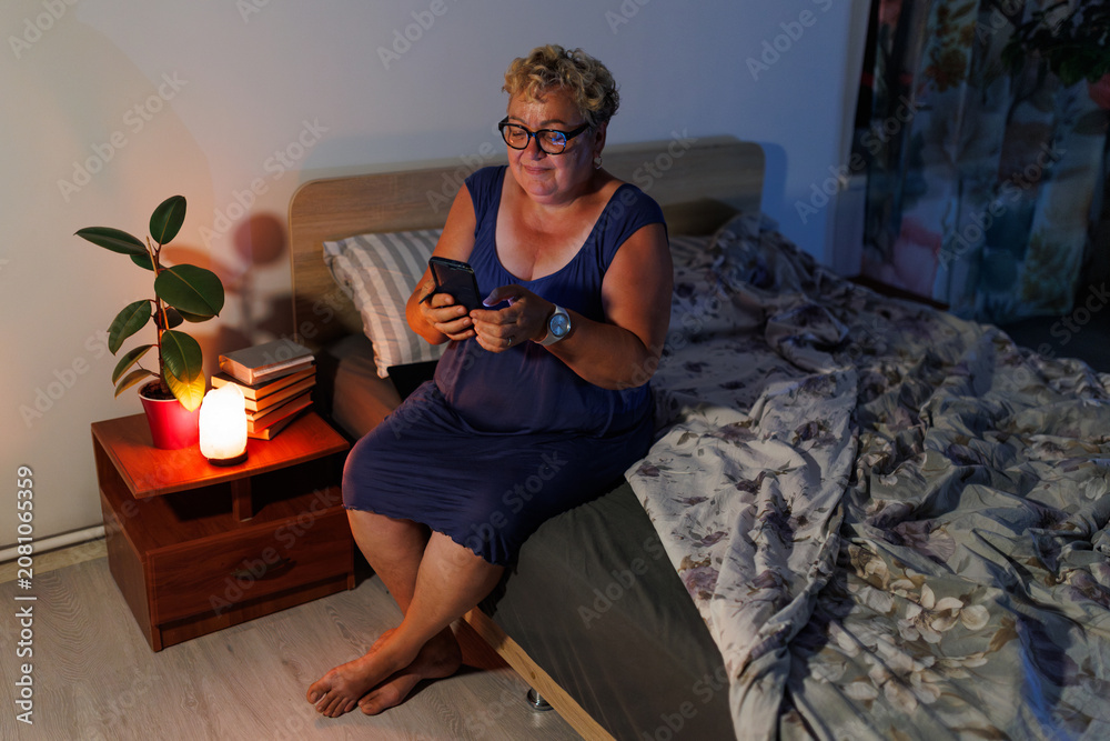 Obraz premium Senior female reading messages on smartphone in dimly illuminated bedroom.