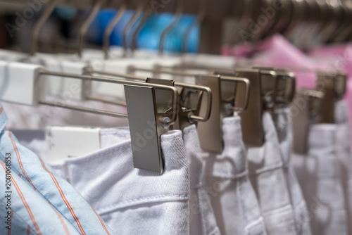 White denim shorts hanging on metal clips in a clothing store, with a blue striped shirt and other garments visible in the background