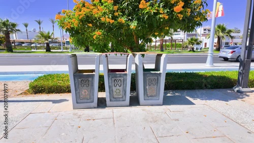 Outdoor recycling bins beneath a yellow flowering tree in Sharm El Sheikh Egypt with tropical landscaping resort buildings and eco friendly waste separation in sunny surroundings
