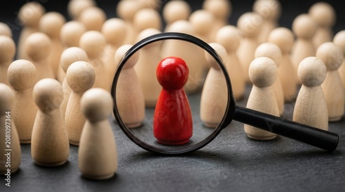 Magnifying glass highlighting a single red figure among many wooden figures to symbolize selection.