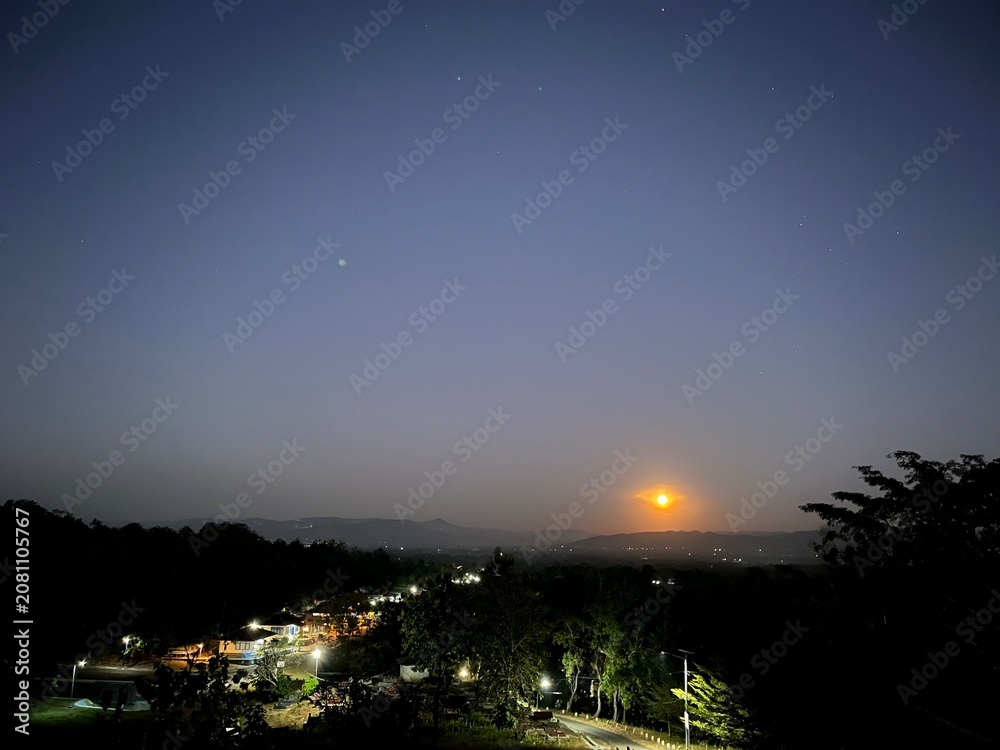 Obraz premium Moonrise over a quiet rural landscape with distant hills, village lights, and a starry evening sky viewed from an elevated viewpoint in a peaceful, inspiring atmosphere.