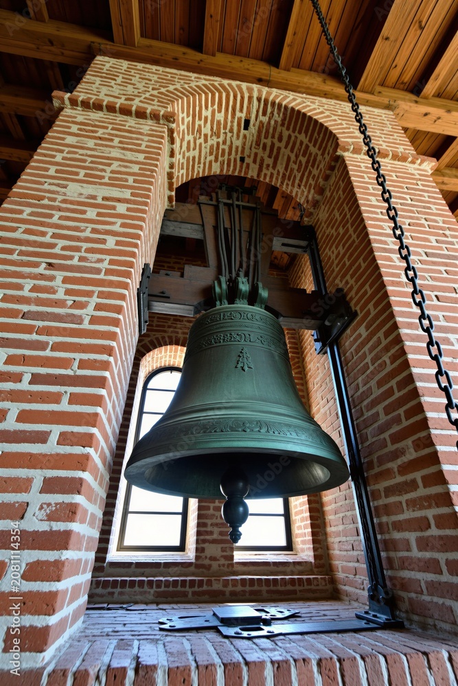 Obraz premium Giant bell hangs inside old brick bell tower. Wooden beams support the structure near arched window. Metal chain attached to bell mechanism.