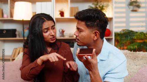 Indian couple man and woman sitting on home sofa argue over smartphone call with angry reaction. Young family man and woman sitting on home sofa complain to operator or delivery service with stress.