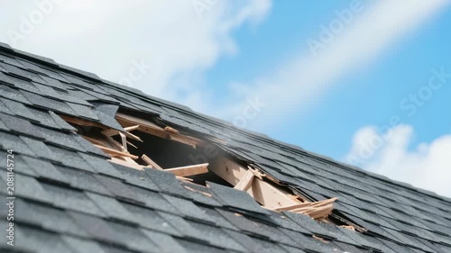 Hole in a residential roof with missing asphalt shingles and exposed wooden rafters against a blue sky, representing storm damage repair or home maintenance.