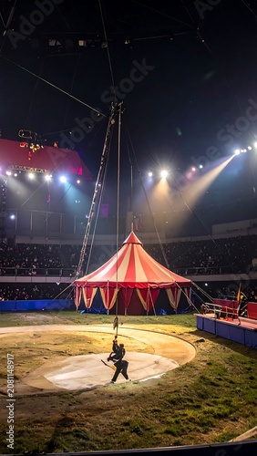 A lone performer swings under a vibrant red and yellow circus tent at night