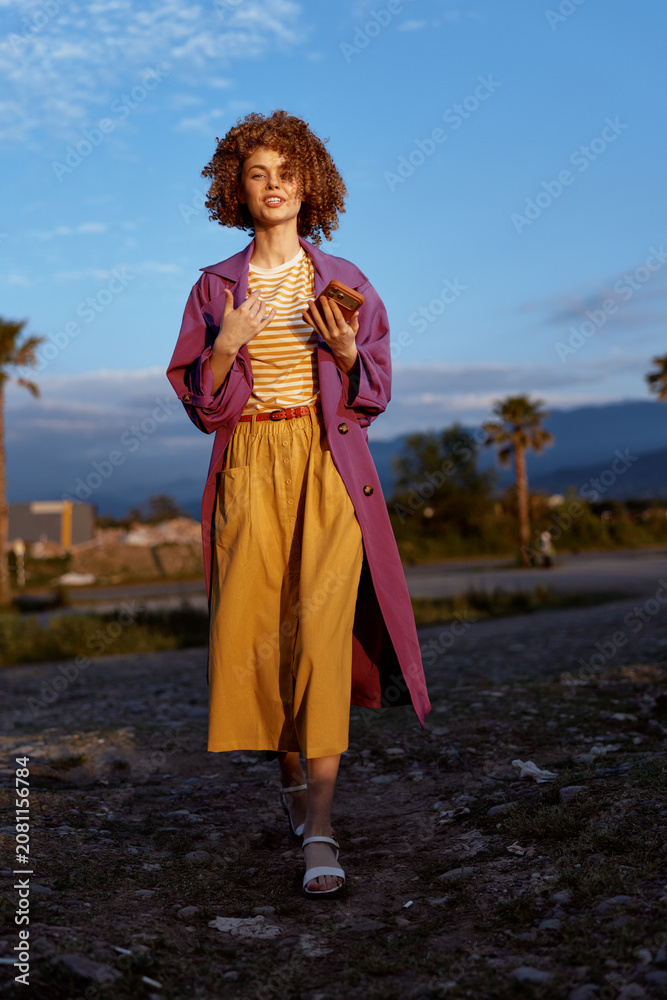 Fototapeta premium Vivid woman outdoors in colorful fashion, striped top, purple coat and mustard skirt, relaxed pose, sunlit scene, natural landscape, lifestyle portrait with bold styling