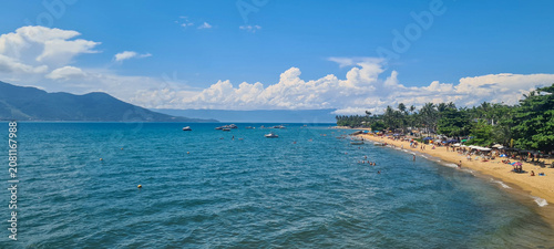 Canvas Print Panoramic view of a beautiful tropical beach with clear blue water, white sand, tourists relaxing, and lush green mountains in the background under a sunny summer sky