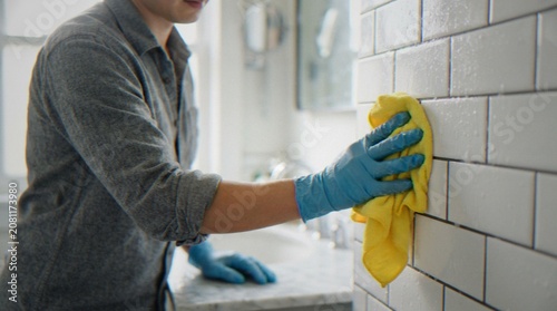 Tableau sur toile A man wearing blue rubber gloves cleaning white subway tiles in a bathroom with a yellow microfiber cloth