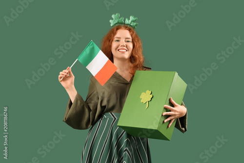 Young redhead woman with gift box and Irish flag on green background. St. Pat...