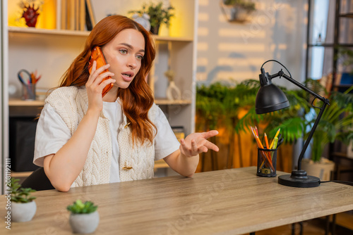 Young woman on home table talks on smartphone explaining details and reacting with hand gesture during call. Adult girl on home table discusses plans with focus and emotion during indoor conversation.