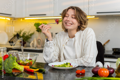 Konstfotografi Young woman eats fresh salad with smile during healthy diet lunch in home kitchen