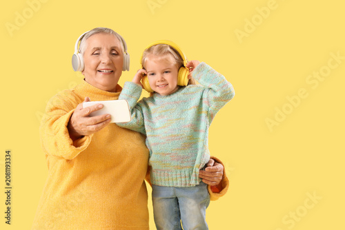 Cute little girl with her granny in headphones taking selfie on yellow backgr...