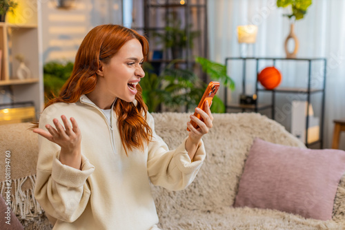 Fotografie Young woman on home sofa talks on smartphone speaker and waves hand in furious dispute showing frustration