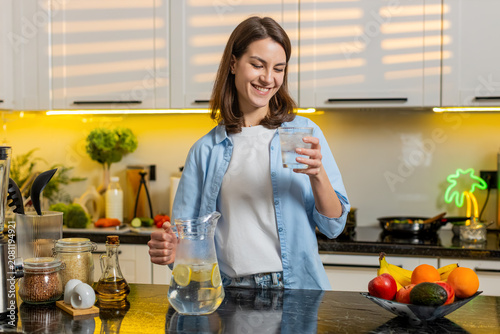 Foto Young woman pours cold lemon water into glass then drinks to quench thirst with happy smile in clean kitchen post workout