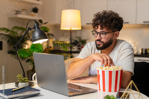 Obraz na płótnie Arabian man at home watches trailer on laptop holds popcorn laughs out loud enjoys preview