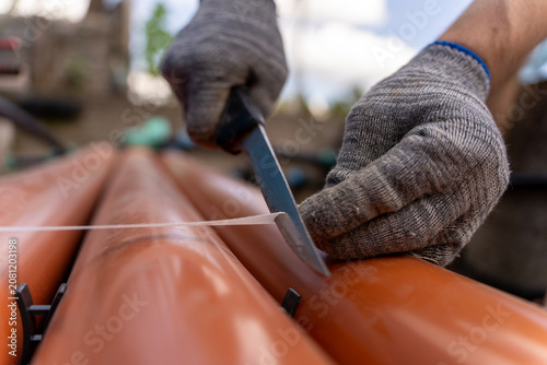 Close up of a worker hand in a protective glove using a knife to cut a plastic strapping band on orange sewage pipes on a construction site during daytime.