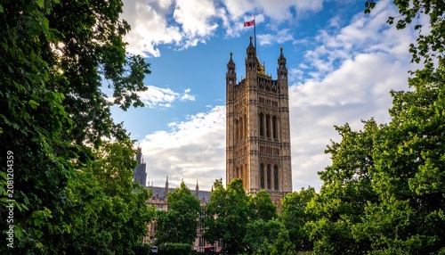 A majestic Gothic-style clock tower rises above lush green trees against a blue sky with white clouds