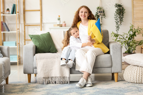 Happy little girl and her mother with Ukrainian flag sitting on sofa in livin...