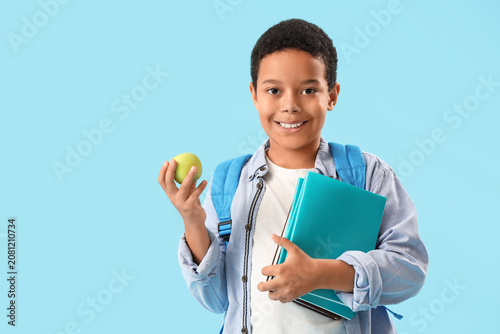 Little African-American schoolboy with books and apple on light blue background
