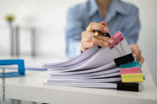 Professional woman organizes documents with colorful clips. She holds a marker, ready to annotate papers. Stacked sheets and vibrant clips add visual interest. Focused demeanor suggests productivity
