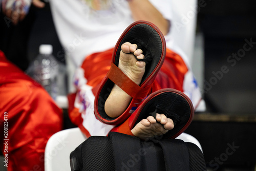Close-up of a teenage kickboxer sitting in a relaxed position with legs crossed, showing his feet wearing protective gear. The athlete is resting between training or competition rounds, highlighting 