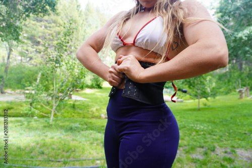 Obraz na plátně A low-angle shot of a sporty woman in a bikini top and leggings adjusting and tightening a black corset waist trainer belt around her midriff with green trees in the background