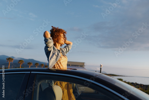 Bild auf Leinwand A young woman leans out of a car window, hair blowing in the wind against a coastal sunset, conveying freedom and carefree summer energy